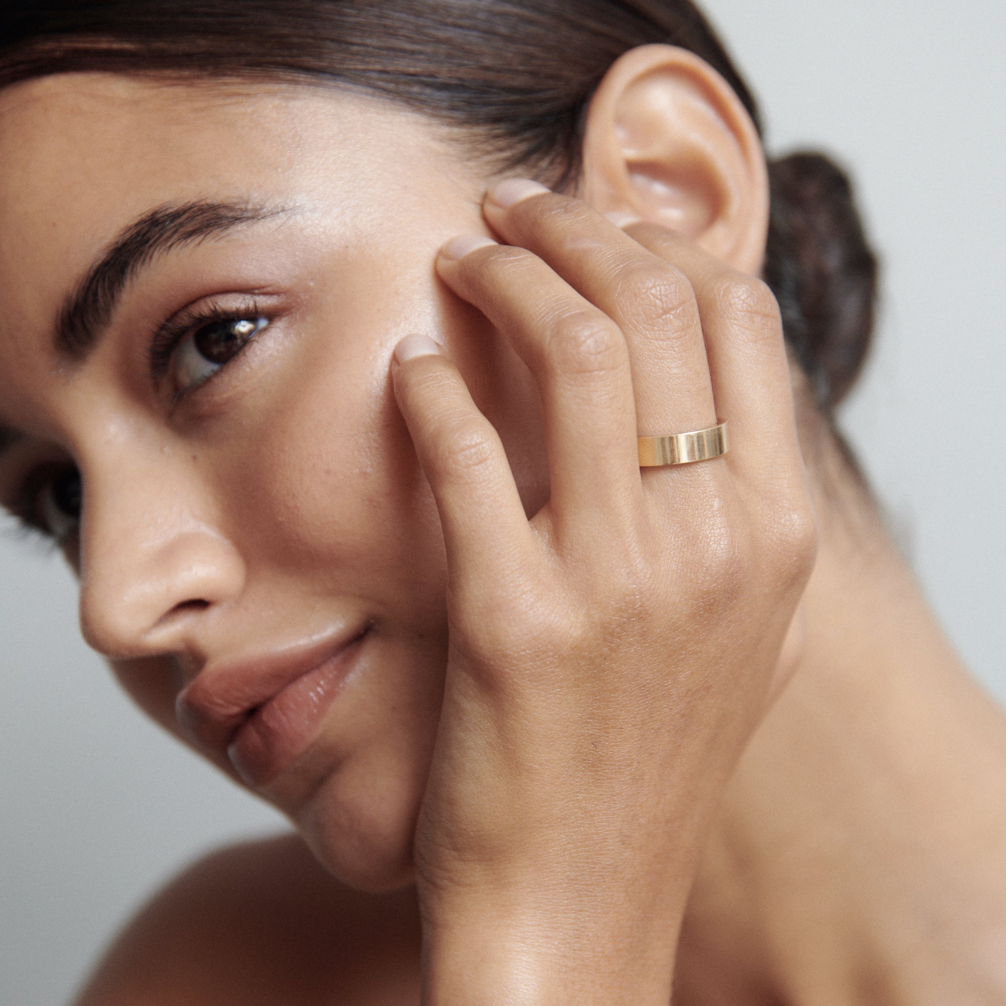 Woman wearing a sleek gold ring, touching her face. The ring stands out against her smooth complexion, highlighting luxury jewellery elegance.  [text overlay: Bold]