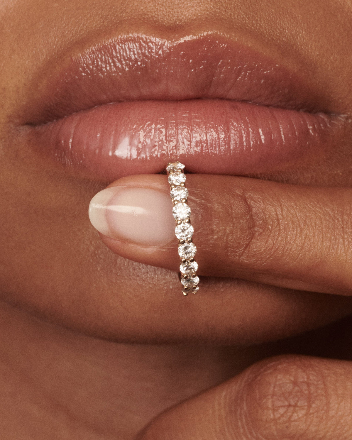 A close-up of a person's lips and fingers wearing a thin gold band ring set with clear round-cut diamond. [text overlay: Francielle wears the 3mm Arraya]