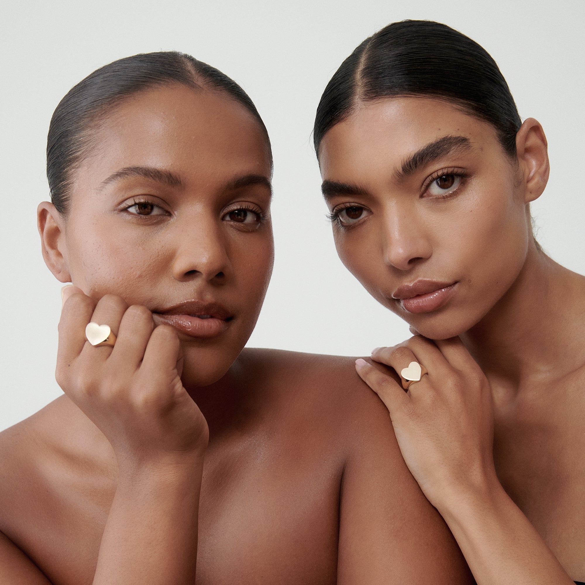 Two women with sleek hair showcase luxury gold heart rings against a neutral background, embodying elegance and minimalism in high-end jewellery.