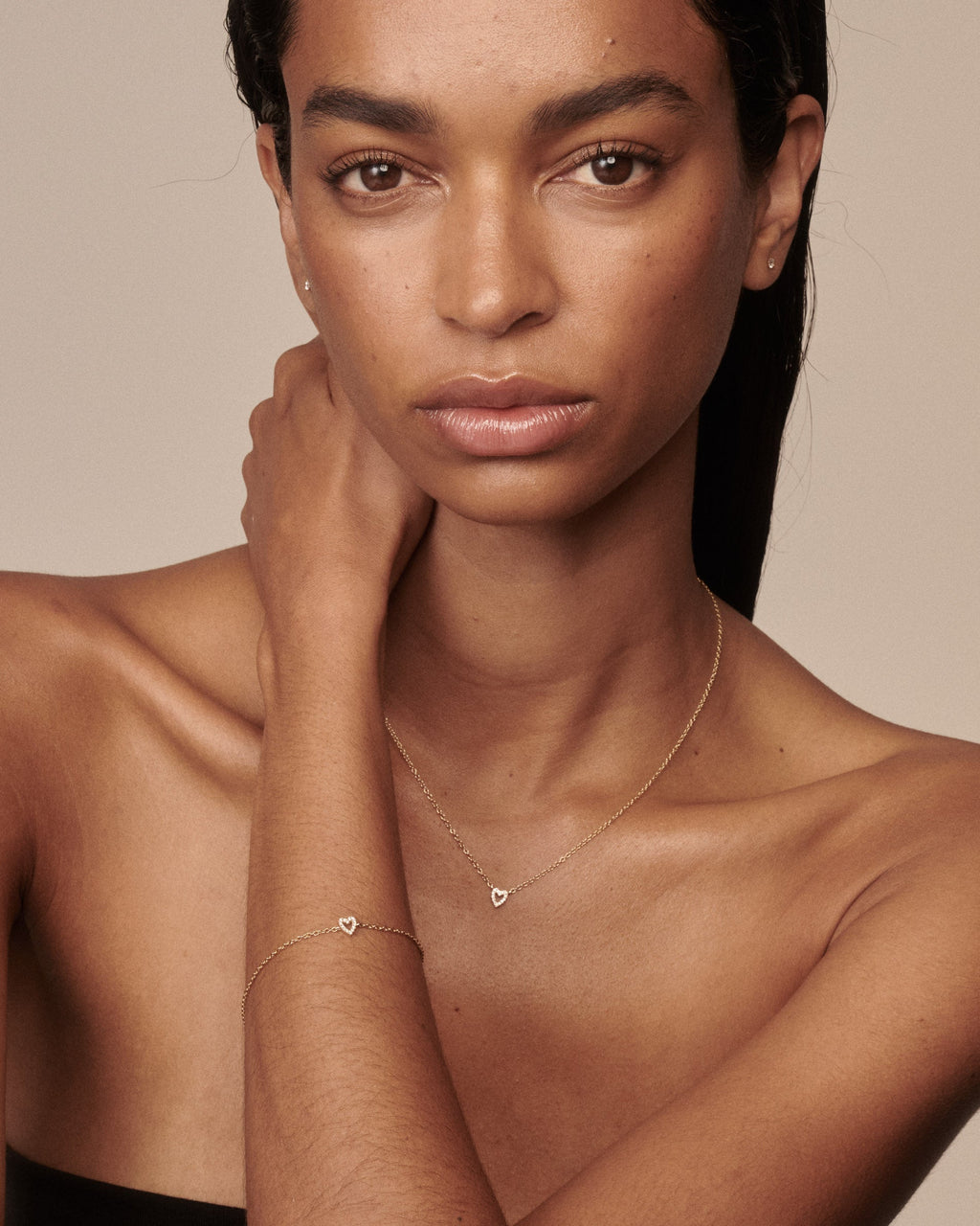 A close-up portrait of a woman wearing delicate gold jewelry, including a thin chain necklace with a small heart pendant and a matching bracelet with a small heart charm, against a warm beige background.
[text overlay: Francielle wears the matching Diamond Heart Necklace & Bracelet]