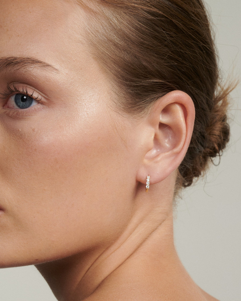 Close-up of a woman's ear wearing a diamond earring against a neutral background [text overlay: Tiny Diamond Huggies]