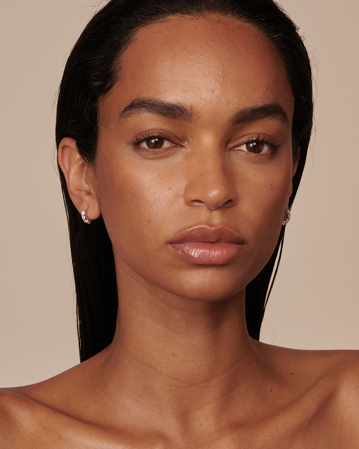A close-up portrait of a woman with dark hair pulled back, wearing small silver hoop earrings against a solid beige background.