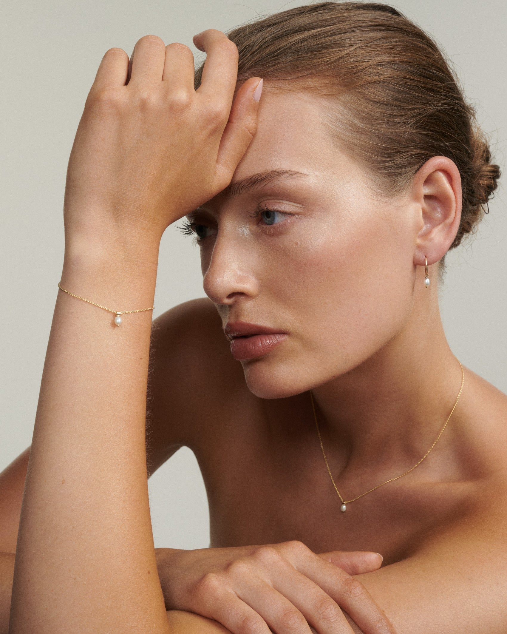 Woman wearing gold jewelry with a plain background [text overlay: Andrea wears the Tiny Pearl Necklace, Bracelet & Hoops]