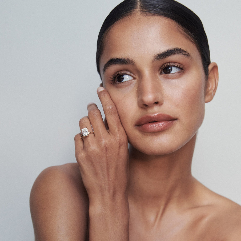 Woman gazes sideways, showcasing a luxury diamond ring on her finger. She has smooth skin, sleek black hair, and wears minimal makeup. [text overlay: Model Wears 2.5ct Diamond]