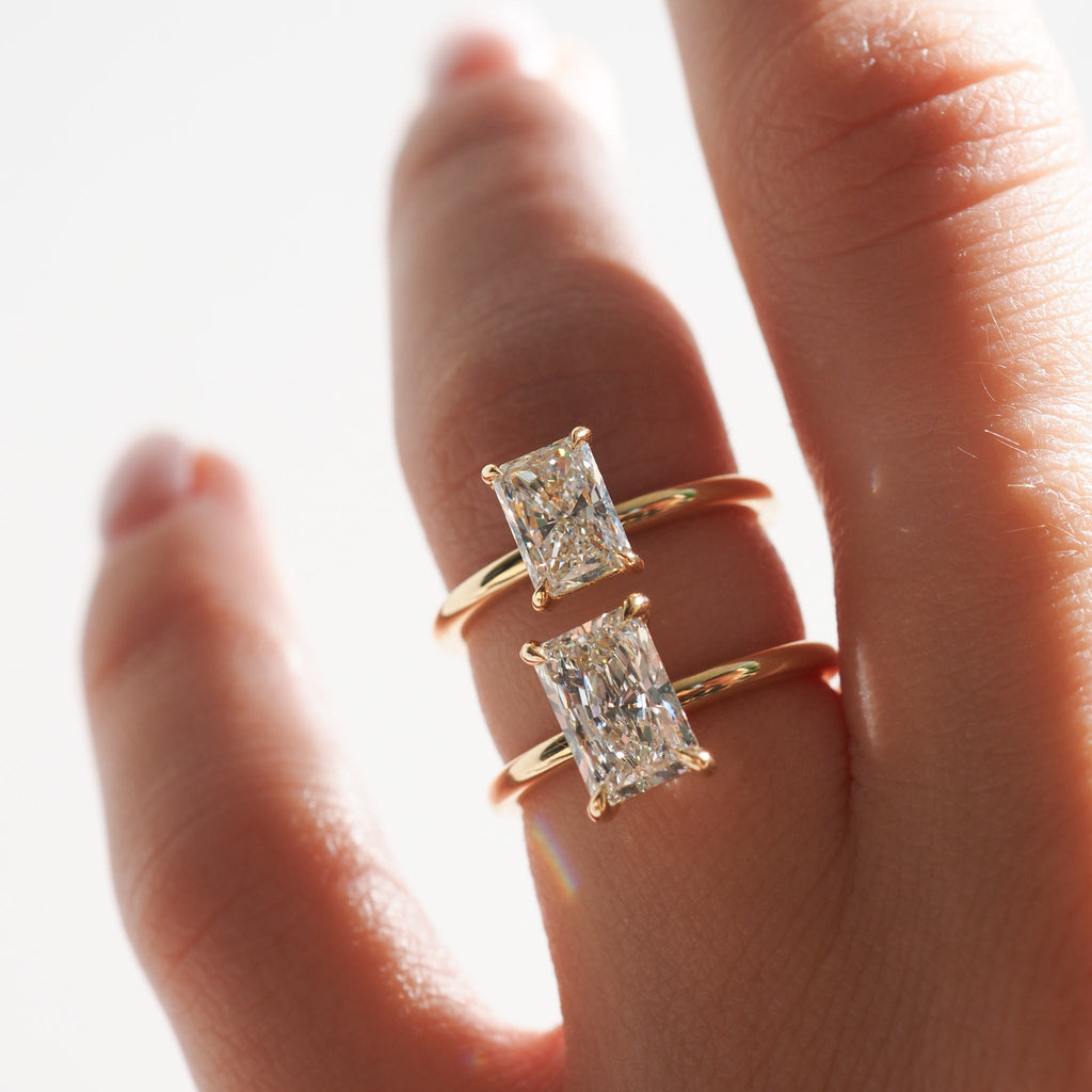 Two gold rings with radiant-cut diamonds displayed on a hand, showcasing luxury jewellery design in natural light.[text overlay: 1ct Diamond (top) and 1.5ct Diamond (bottom)][text colour: #000000]